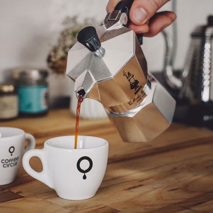 Bialetti Moka Express - Espressokocher gießt frisch gebrühten Kaffee in eine weiße Tasse auf Holzuntergrund.